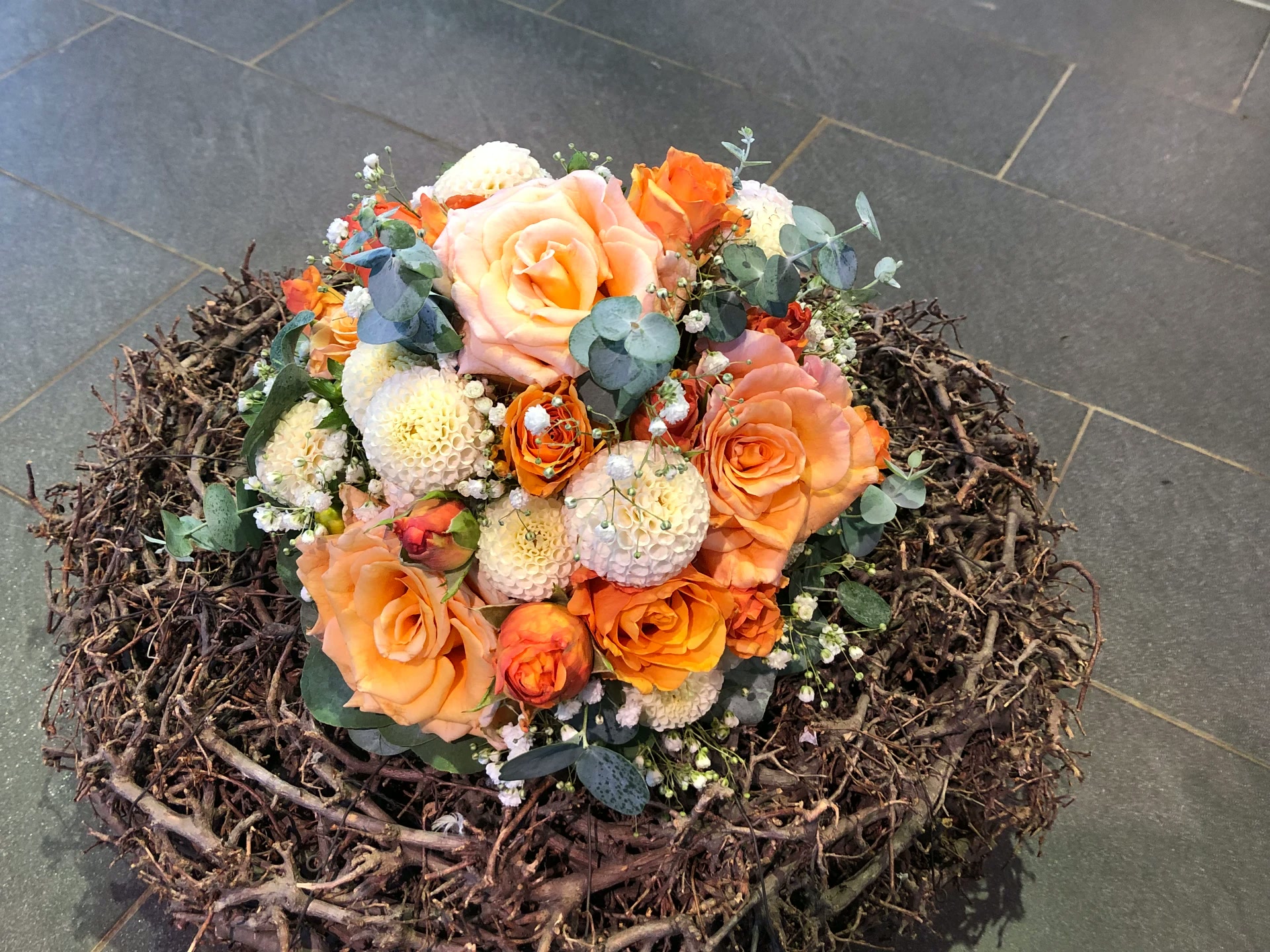 Rustikales Gesteck mit orangefarbenen Rosen, weißen Ballchrysanthemen und Eukalyptus auf einem Rebenkranz