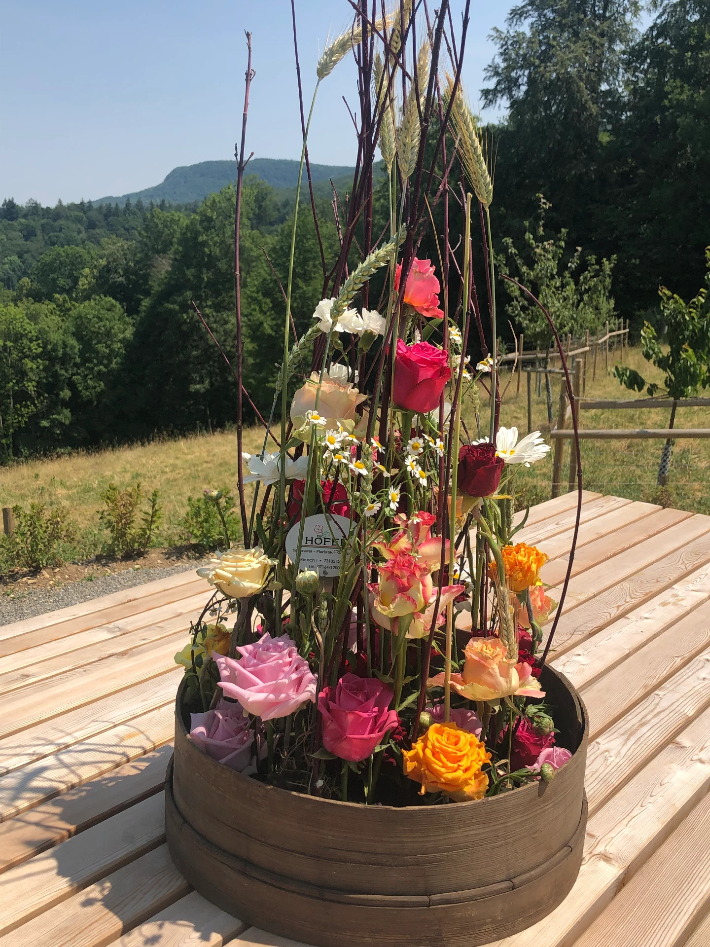 Üppiges Sommergesteck mit Rosen, Margeriten und Getreidehalmen in einer Holzschale vor Landschaftskulisse