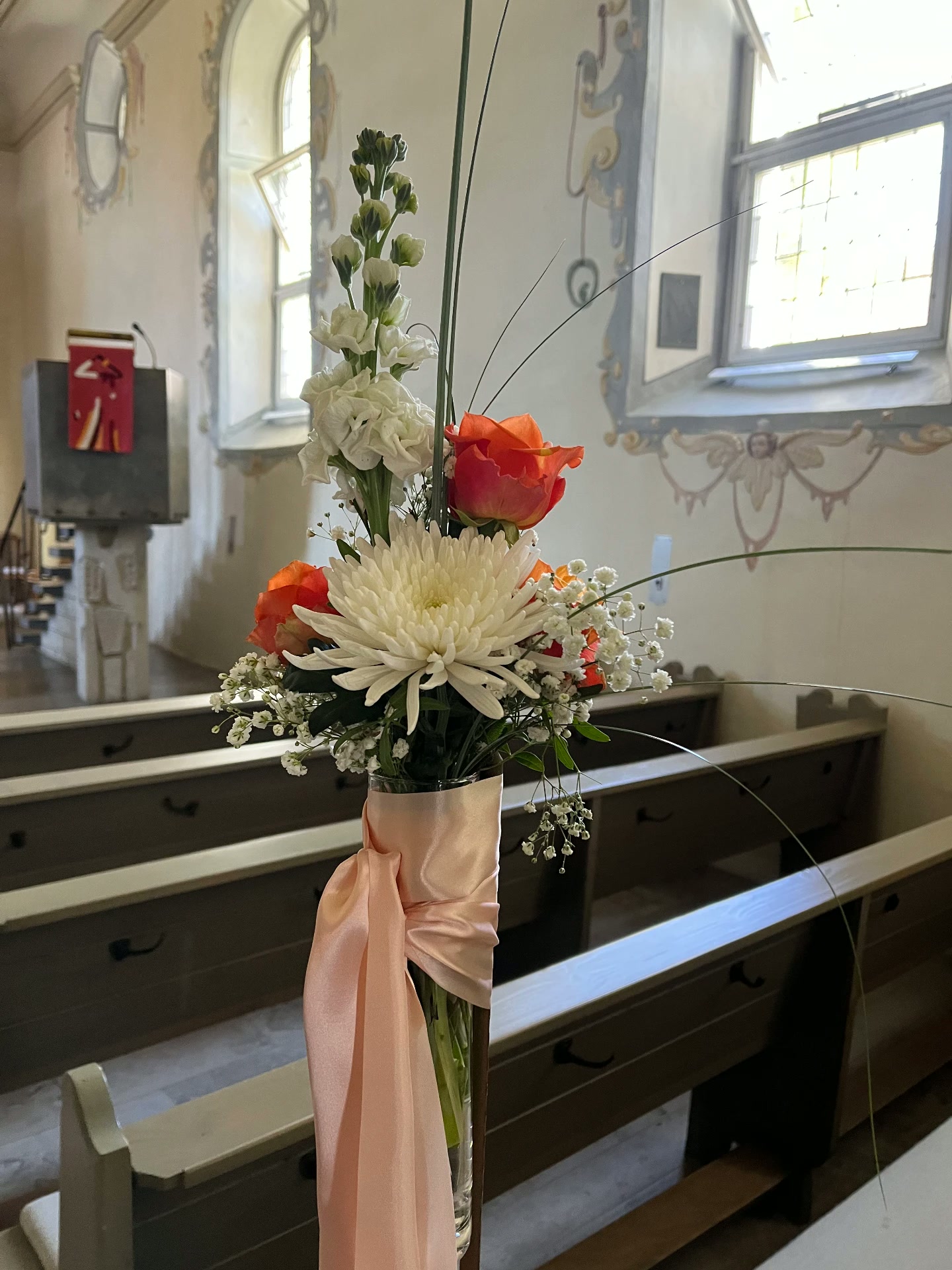 Blumengesteck mit weißer Chrysantheme, Rose und Levkojen mit rosa Schleier an einer Kirchenbank