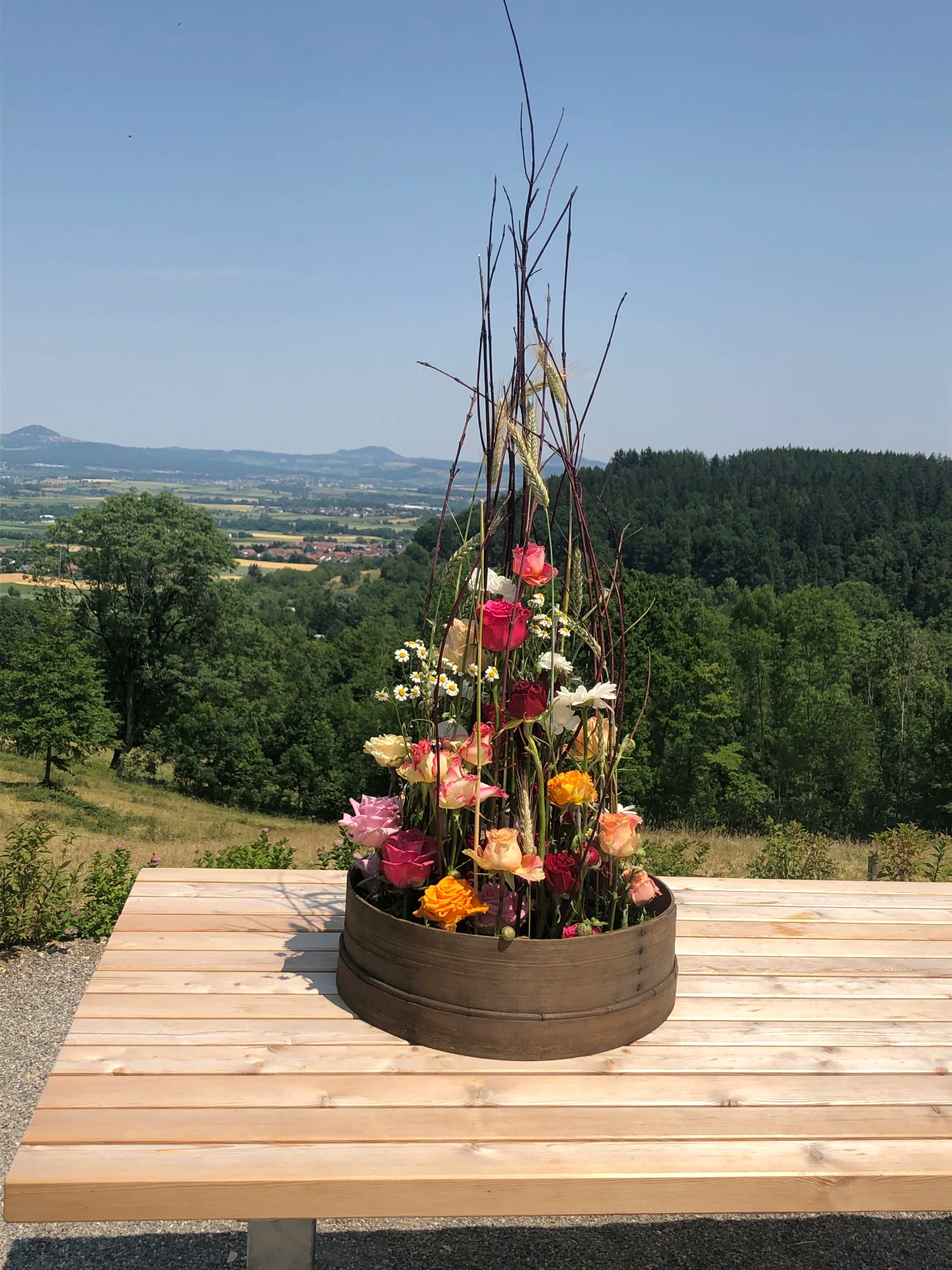 Hohes Gesteck mit Rosen, Margeriten und Ähren in einer Holzschale auf einer Festtafel mit Weitblick